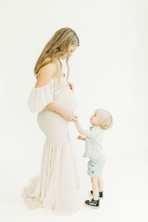 A toddler in light blue clothing reaches up to touch a woman wearing a flowing cream-colored dress against a white backdrop.