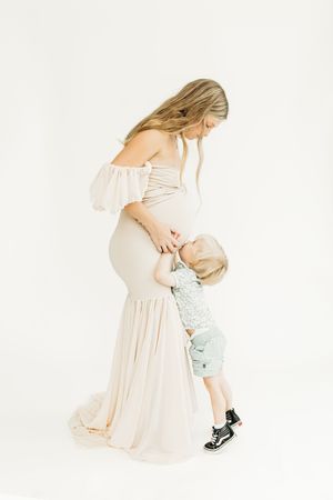 Mother in soft peach dress embraces small child wearing light blue outfit against white background.