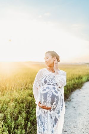 Woman in flowing white lace dress poses gracefully in a sunlit field beside a winding dirt path at golden hour.