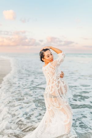 A figure in a white lace dress poses gracefully in ocean waves during sunset creating a dreamy beach portrait.