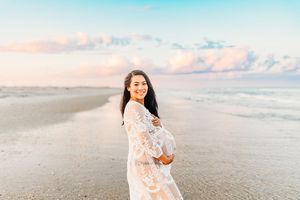 A scenic beach photoshoot at sunset with soft pastel colors and flowing white lace dress against the ocean waves.