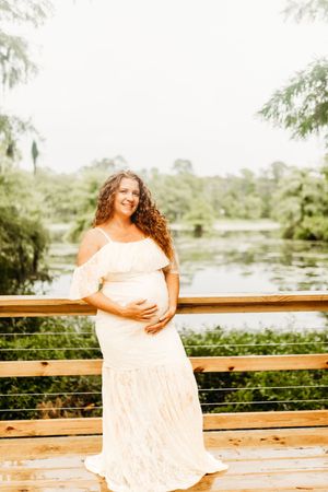 A pregnant woman in a flowing white off-shoulder dress stands on a wooden deck overlooking a scenic lake.
