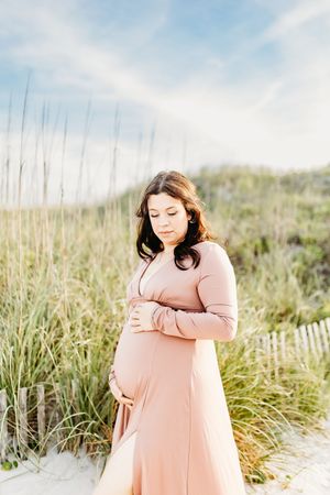 A person in a flowing blush pink dress stands thoughtfully among beach grass on a sunny day.