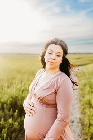 A woman in a pink dress stands in a sunlit field of tall grass while the wind blows her hair.