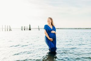 Woman in flowing blue dress stands in calm water during sunset maternity photo session at lake.