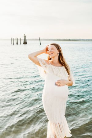 A person in a flowing white dress stands by the water at sunset with wooden posts visible in the background.