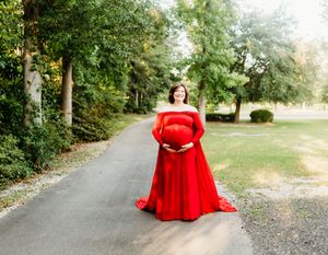 A person in a flowing red dress poses gracefully along a tree-lined path in a park setting.