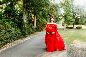 A person in a flowing red dress poses gracefully along a scenic tree-lined path in a natural outdoor setting.