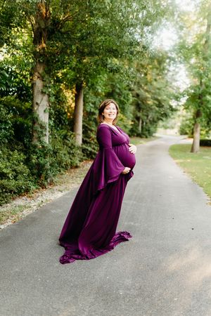 A person in a flowing purple gown twirls gracefully on a tree-lined path during a photo session.