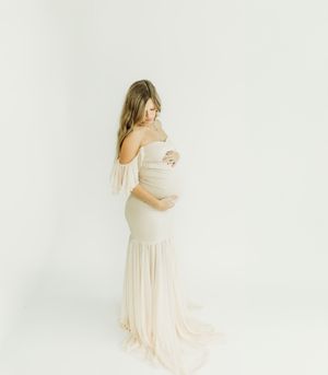 A pregnant woman in a flowing cream dress poses gracefully in a bright white studio setting.