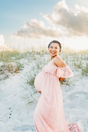 A maternity photo at sunset shows a woman in a flowing pink off-shoulder dress standing on white sand dunes.