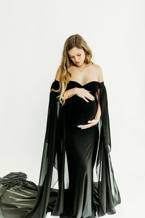 Elegant maternity photoshoot featuring a black off-shoulder gown with flowing cape against white backdrop.