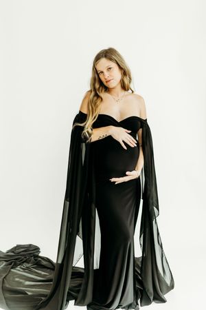 Dramatic maternity portraits with a black off-shoulder gown and sheer cape against white backdrop.
