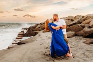 A couple in blue and white sits on rocky coastal beach during golden sunset.