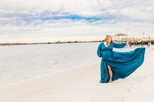 Woman in flowing teal dress twirls gracefully on pristine white sand beach at sunset.