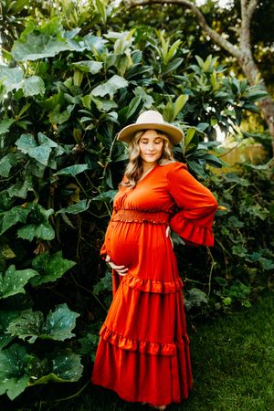 A woman in a flowing orange dress and straw hat poses gracefully in a lush garden setting.