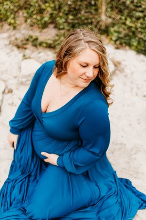A person in a flowing royal blue dress sits gracefully on a sandy beach with greenery in the background.