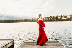 A person in a vibrant red dress stands gracefully on a wooden dock overlooking a serene lake at sunset.