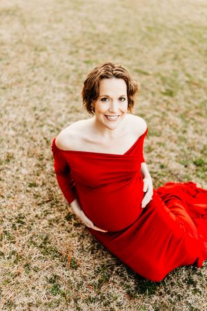 Expectant mother wearing flowing red off-shoulder dress sits gracefully on grass during outdoor maternity photoshoot.