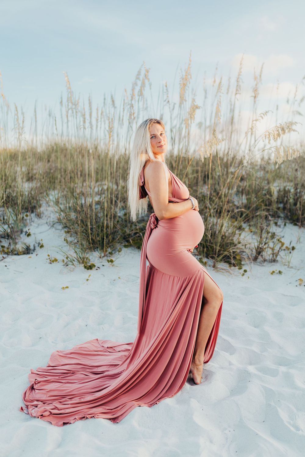 A person in a flowing pink maternity gown poses gracefully on white sand beach with sea oats at sunset.