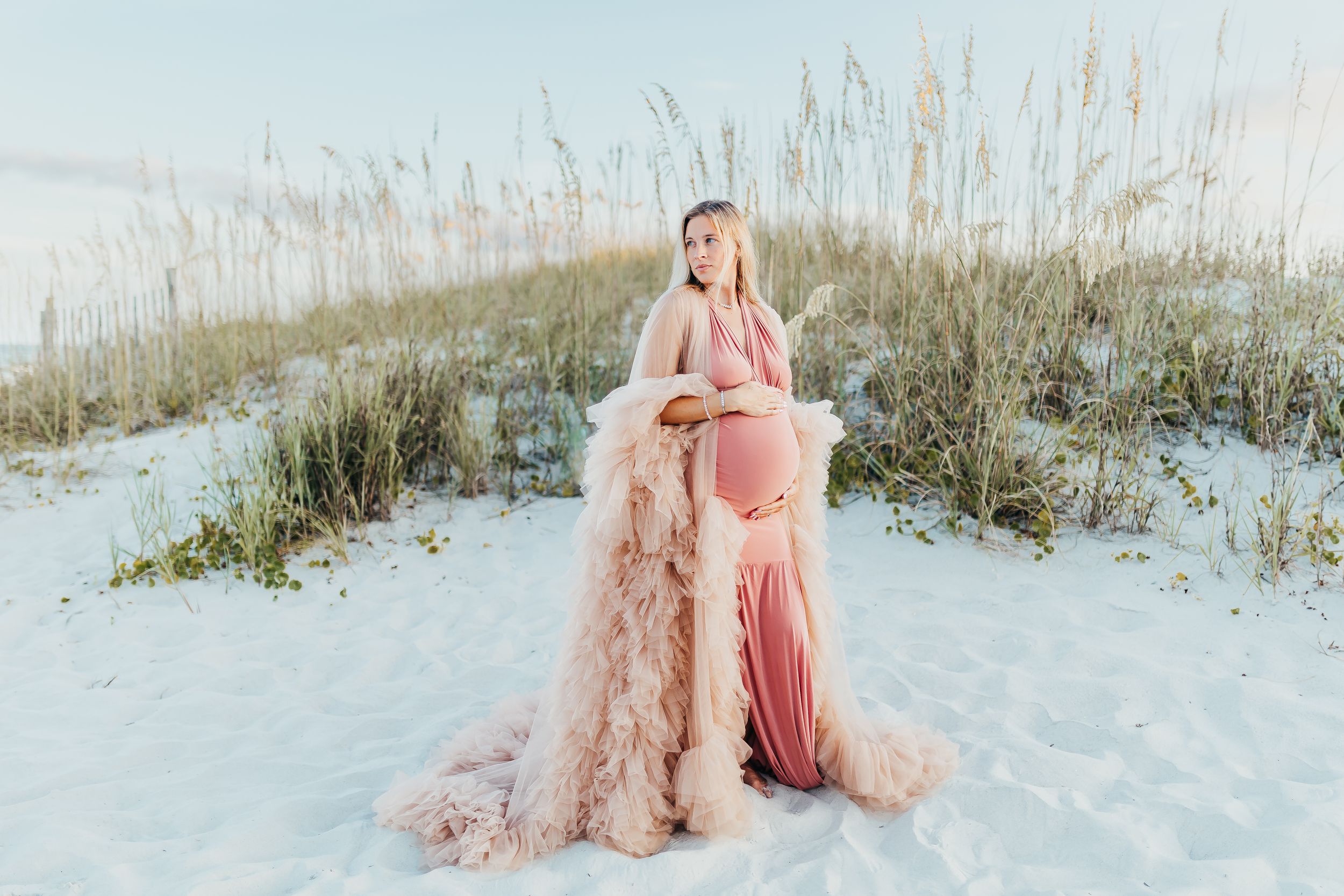 A person in a flowing pink dress stands in snow-covered sand dunes with sea oats at sunset.