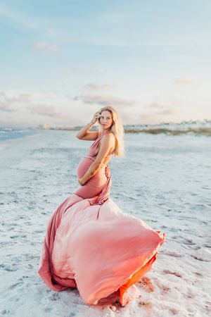A flowing coral pink dress twirls gracefully against white sand and ocean waves at sunset.