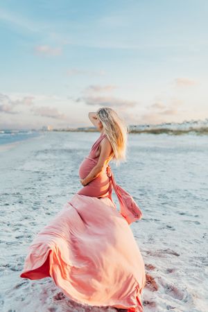 A person in a flowing coral pink dress twirls gracefully on a sandy beach at sunset with ocean waves in the background.