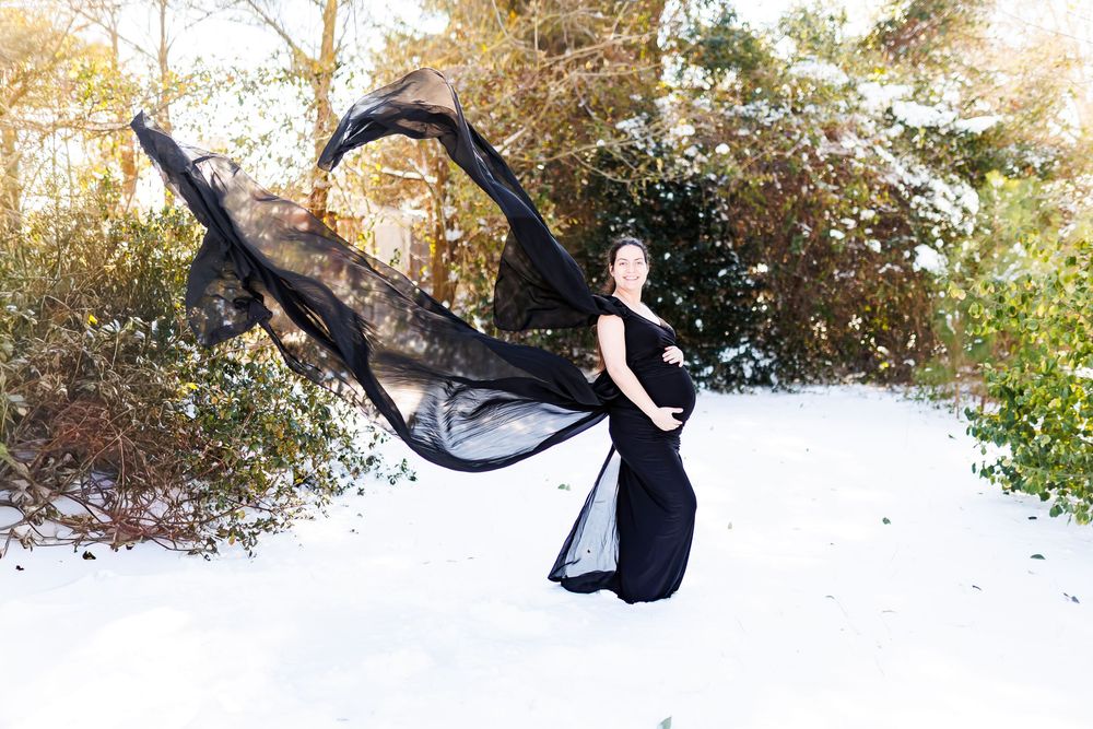 A flowing black dress and sheer fabric billows dramatically against a snowy winter landscape with trees in the background.