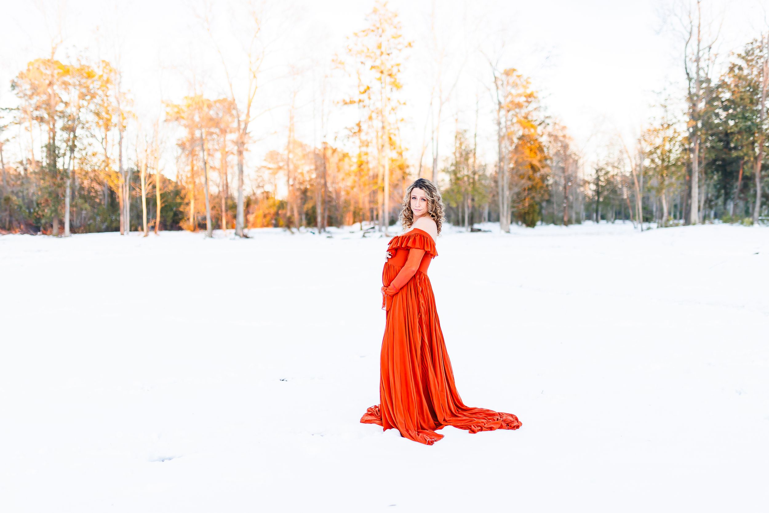 A figure in a flowing red dress stands in a snowy winter landscape during golden hour sunset.