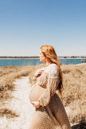 A person in a beige sweater sits in tall golden grass by the beach on a sunny day.