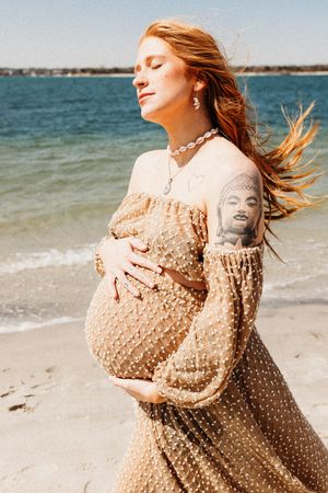 A pregnant woman in a flowing polka dot dress stands on a beach with wind-blown hair and ocean waves behind her.