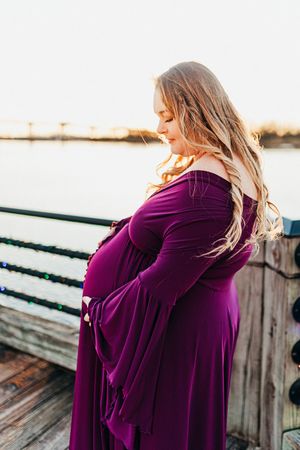 Expectant mother in flowing purple dress stands on wooden dock at sunset with wind blowing through her hair.