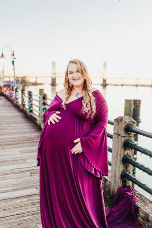 A maternity photoshoot on a wooden pier features a flowing burgundy dress against a bright outdoor setting.