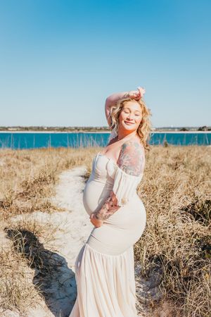 A person in a flowing white dress stands joyfully on a beach with tall grass and blue ocean in the background.