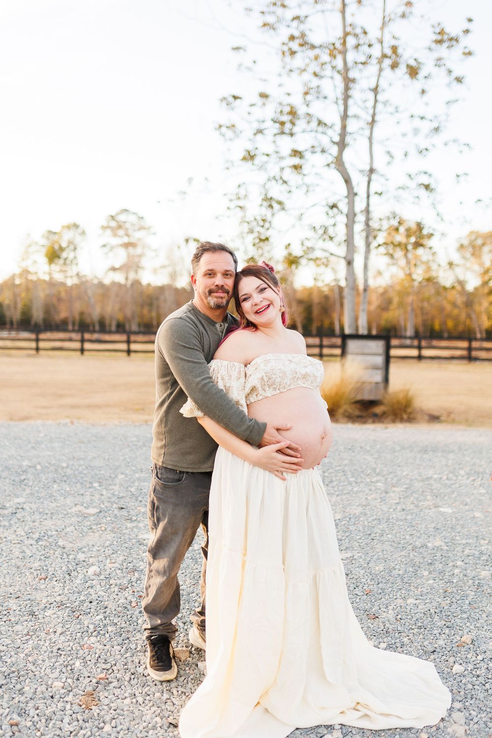 A couple poses outdoors in an autumn setting, with a flowing white maternity dress and casual attire.