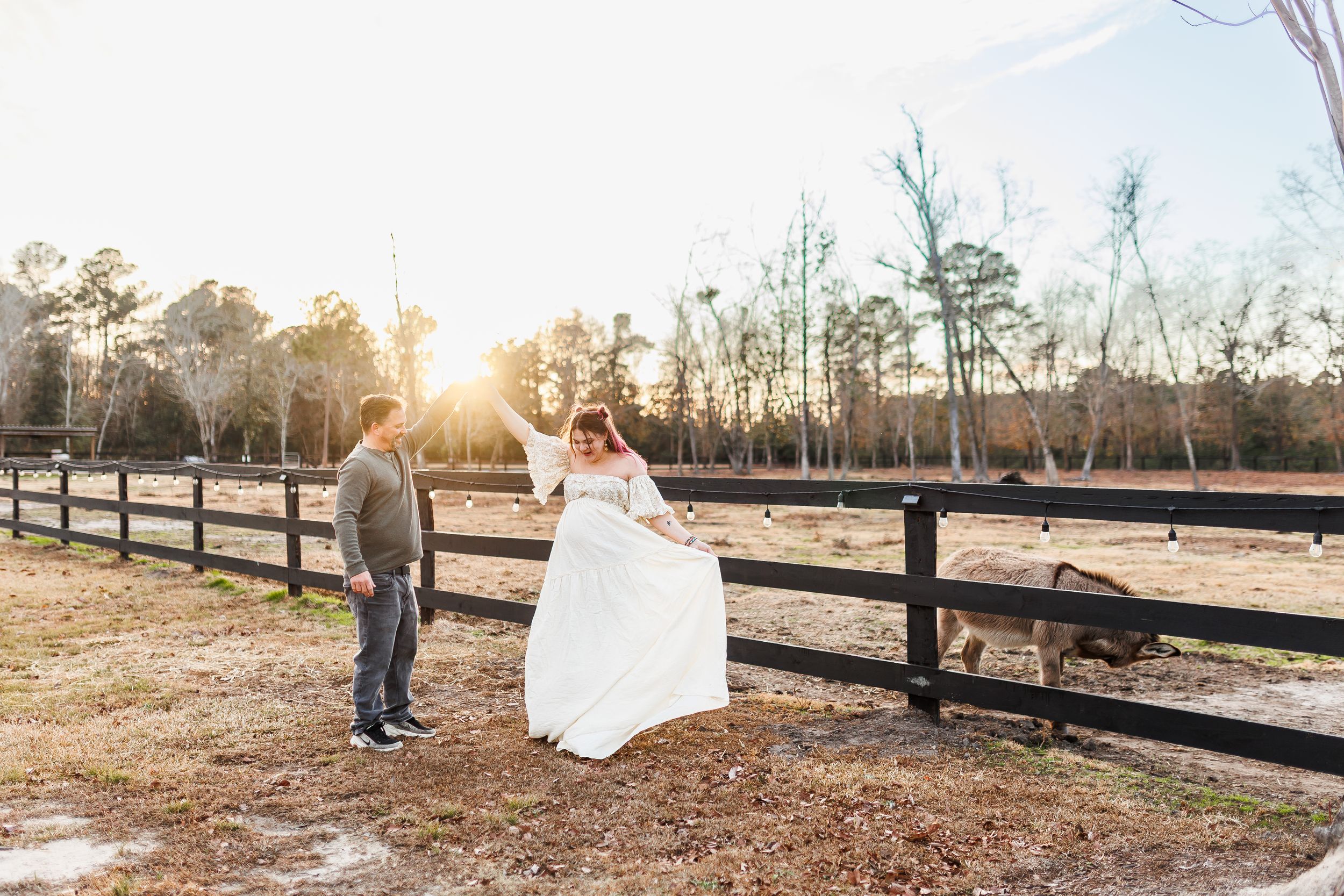 A couple in wedding attire walks through a rustic outdoor setting at sunset with wooden fences in the background.