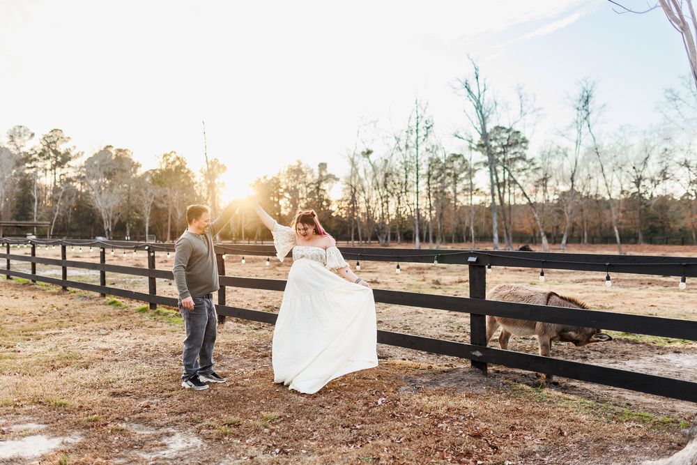 A couple in wedding attire walks through a rustic outdoor setting at sunset with wooden fences in the background.