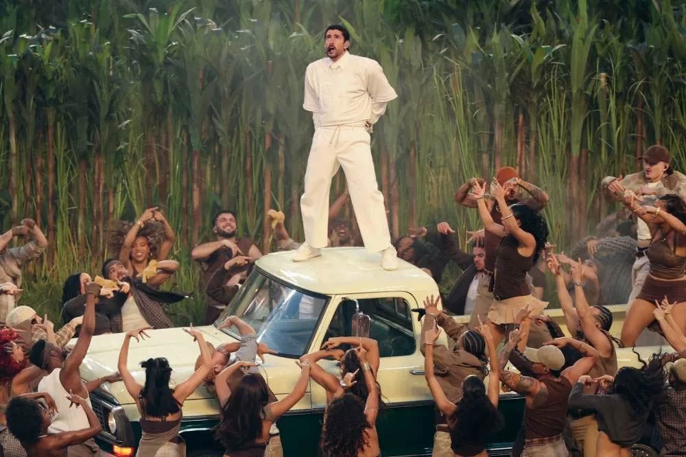 Artist, Bad Bunny, in white stands atop a car surrounded by an enthusiastic crowd in front of tall reeds.