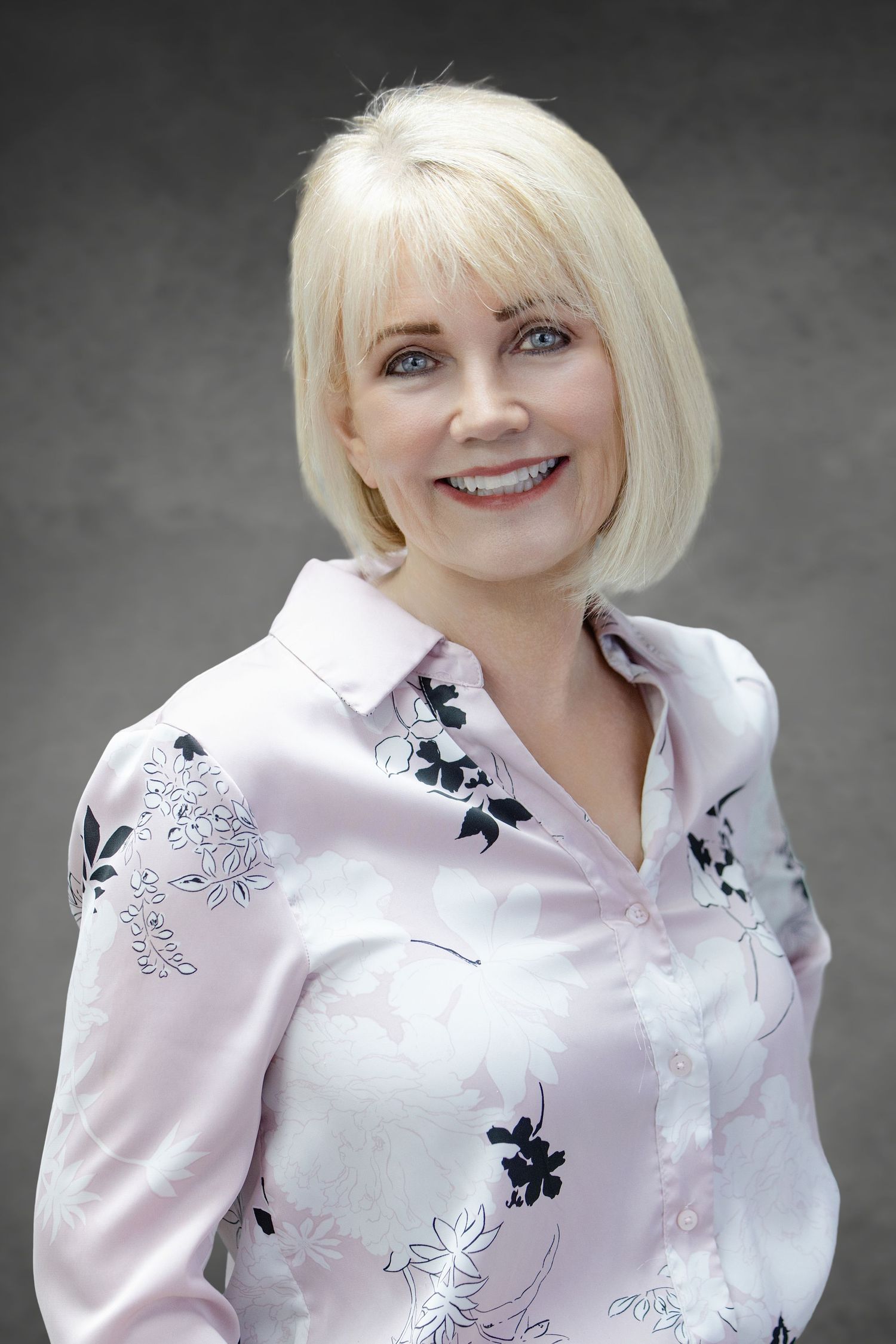 Professional portrait featuring a white blouse with black floral embroidery against a dark gray background.