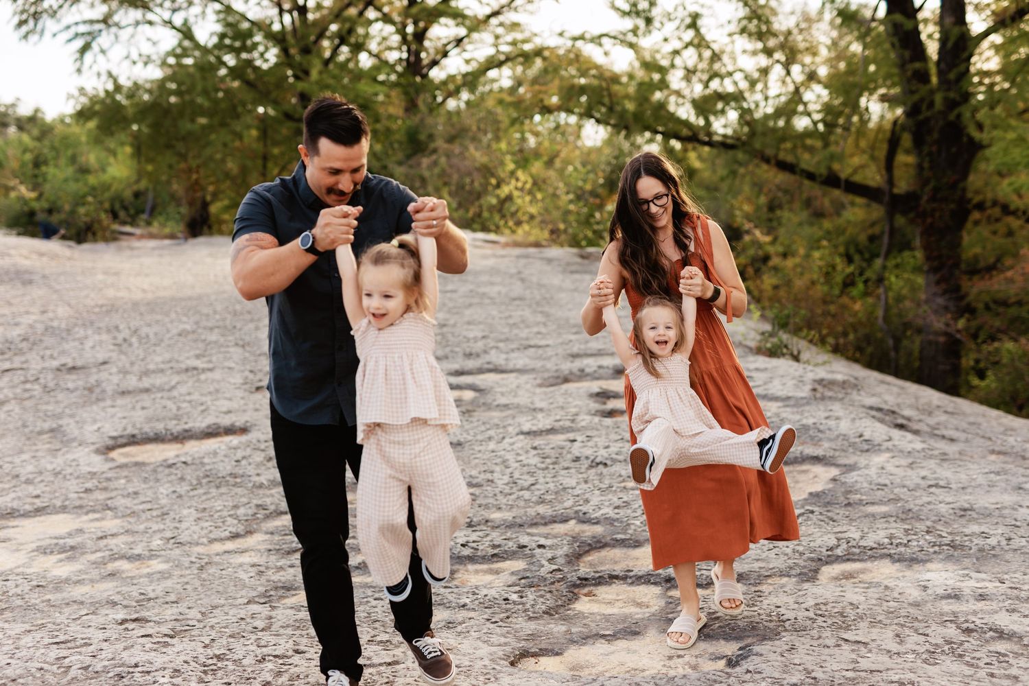 Parents joyfully swing their two young daughters outdoors on rocky terrain surrounded by trees during a family photo session.