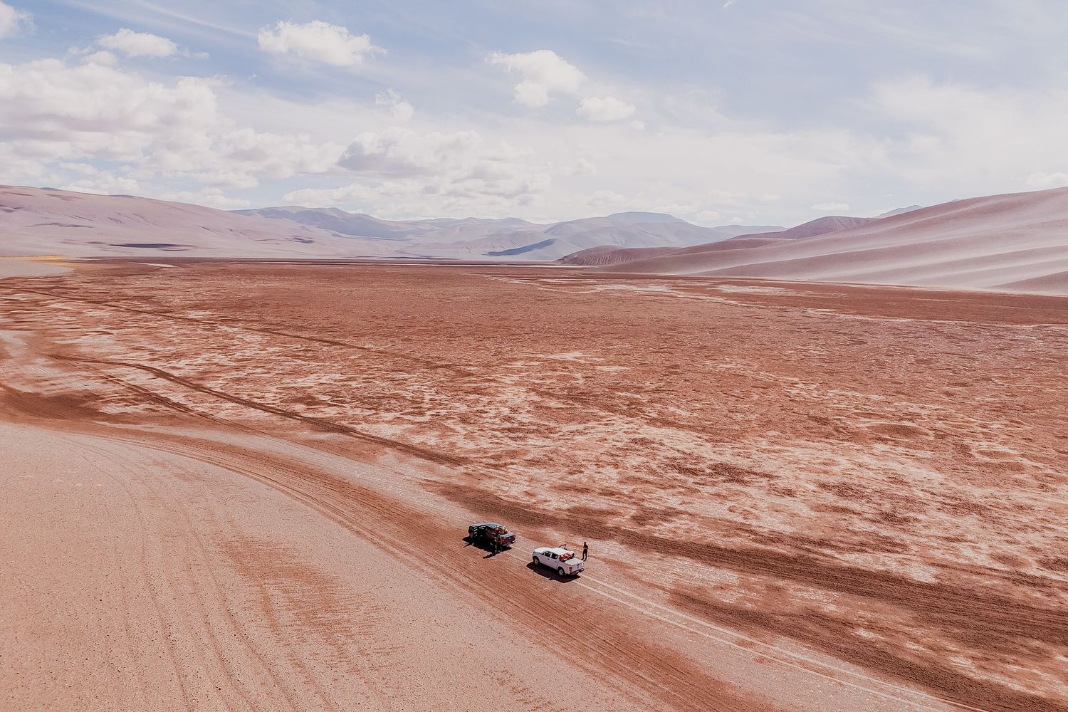 Off-road vehicles traverse a vast desert landscape with rolling red sand dunes stretching to the distant mountains.