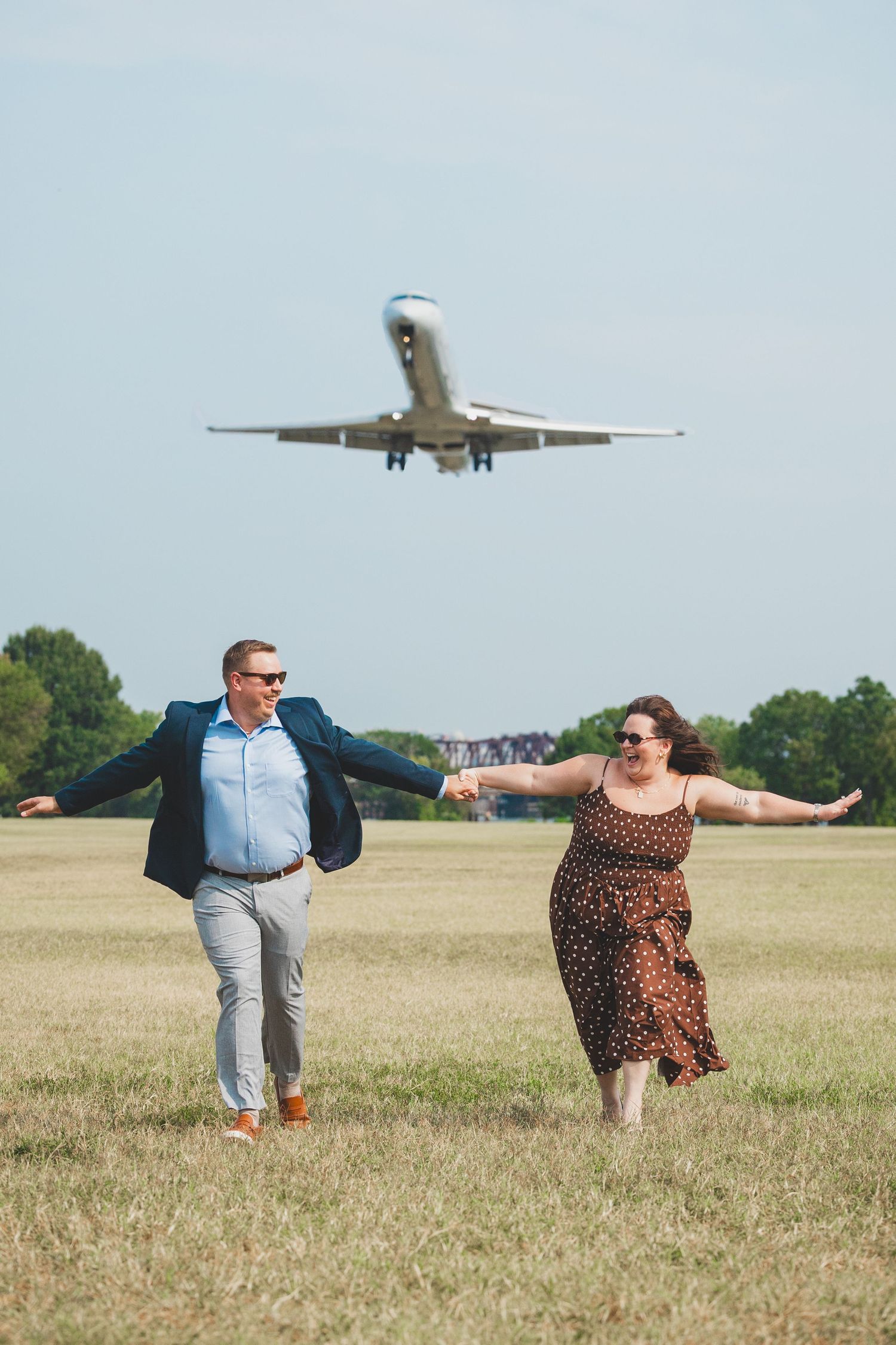 Two people pose playfully in a grassy field as an airplane flies overhead in the background.