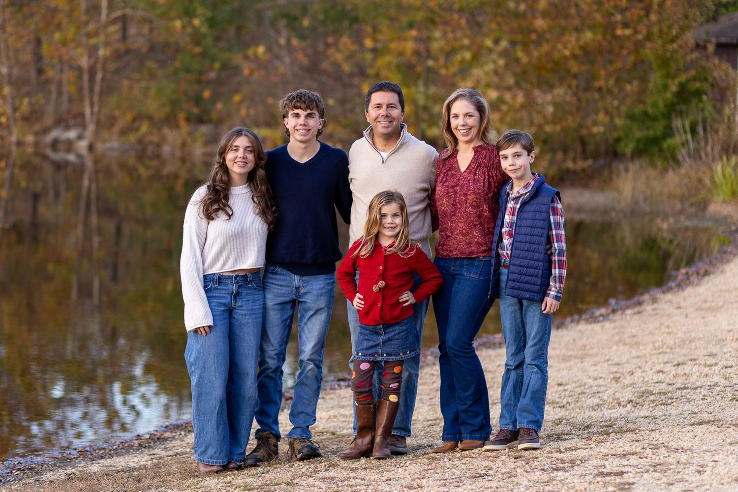 Family group poses together outdoors in front of a lake reflection with fall foliage in background.