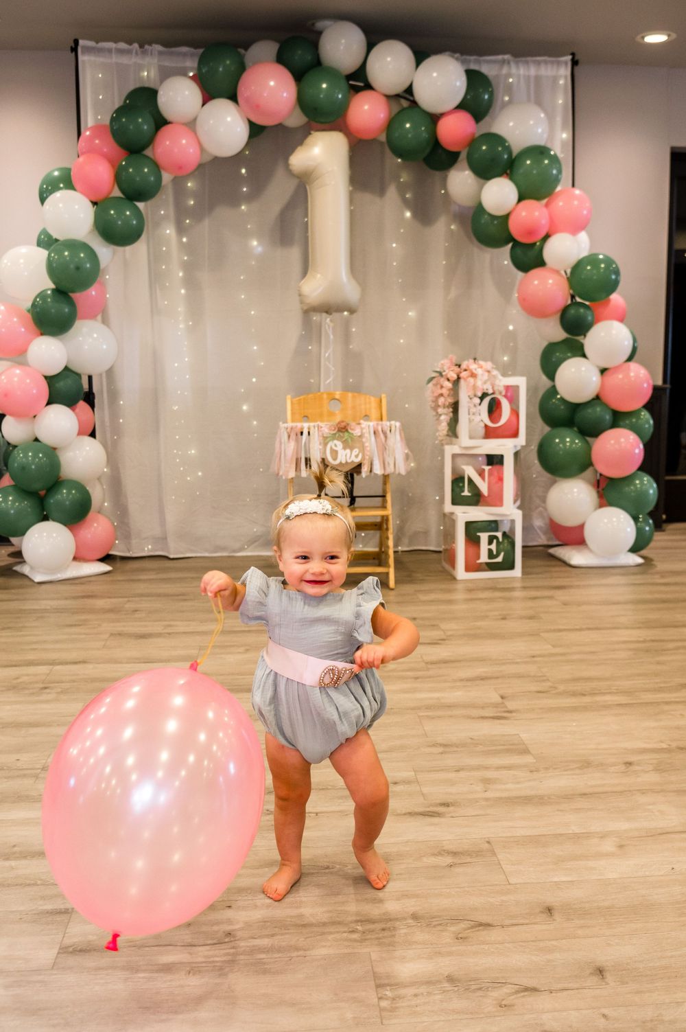 A baby in a gray onesie plays with pink and green balloon decorations during a first birthday celebration.