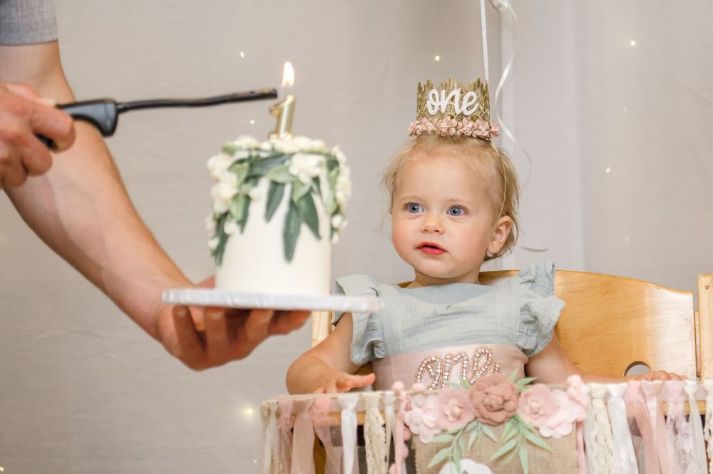 A small child wearing a birthday crown looks on as someone holds a cake with a sparkler at a pink-decorated party.