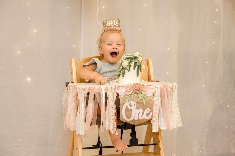 A baby in a high chair celebrates their first birthday with a cake smash photoshoot against a sparkly backdrop.