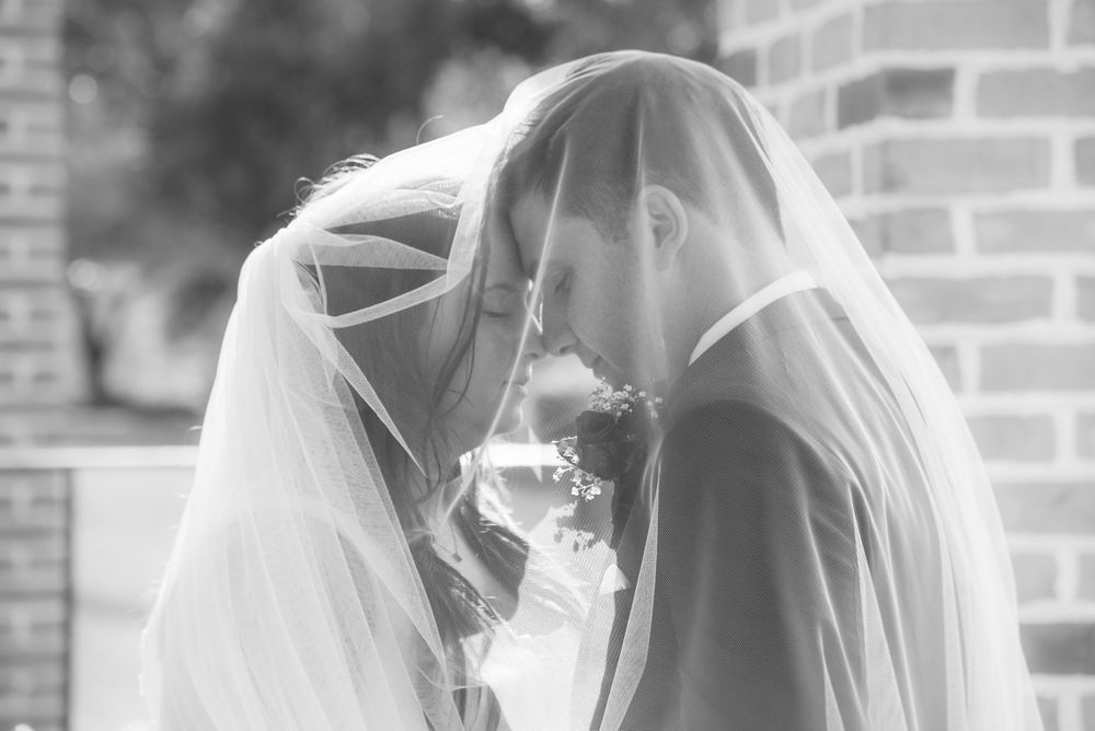 A romantic black and white wedding photo showing a couple sharing an intimate moment under a flowing bridal veil.