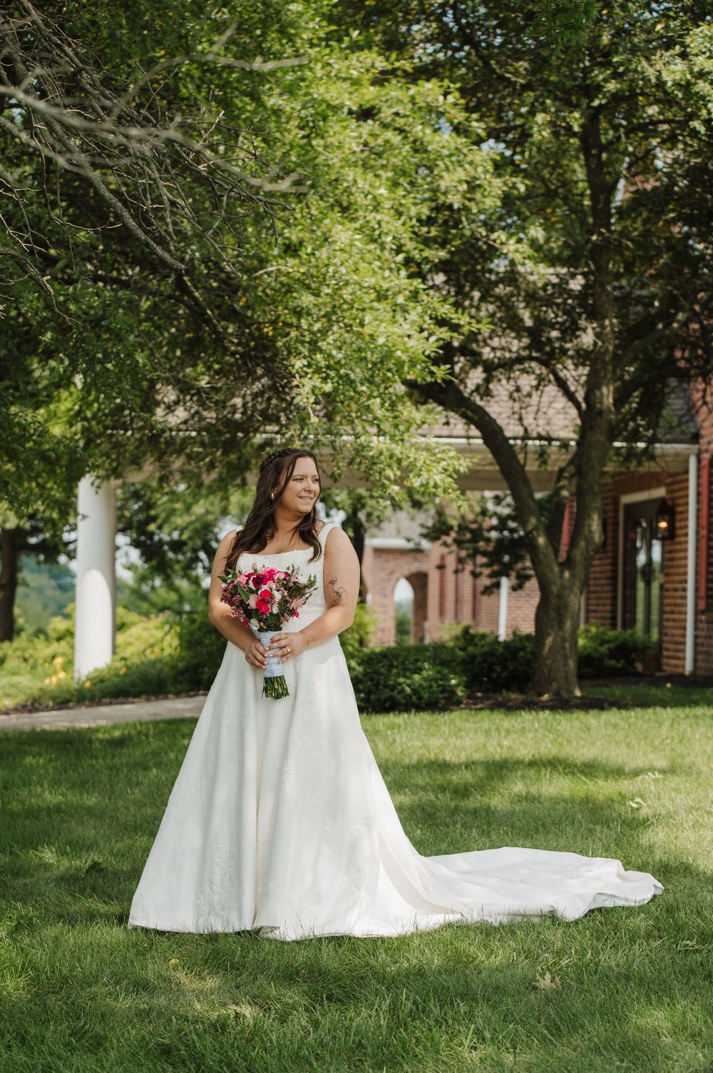 A bride in a flowing white wedding dress poses on a lush green lawn with trees and brick buildings in the background.