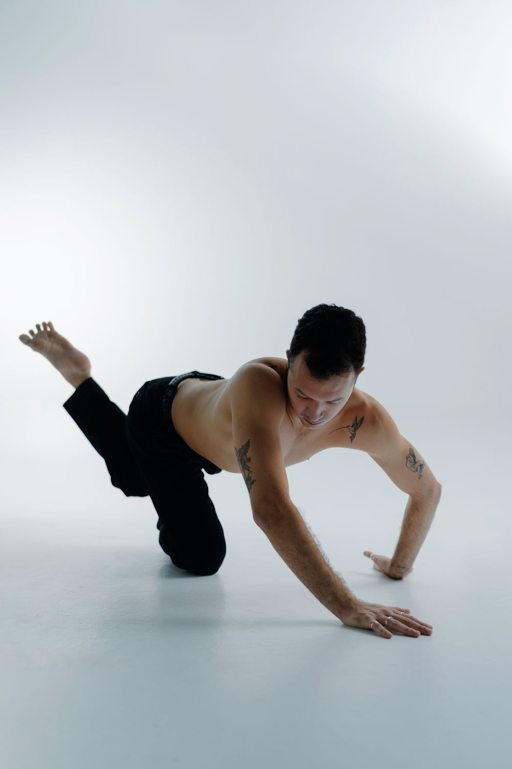 A shirtless dancer performs a contemporary dance move on a light gray background, balancing on one arm and knee.