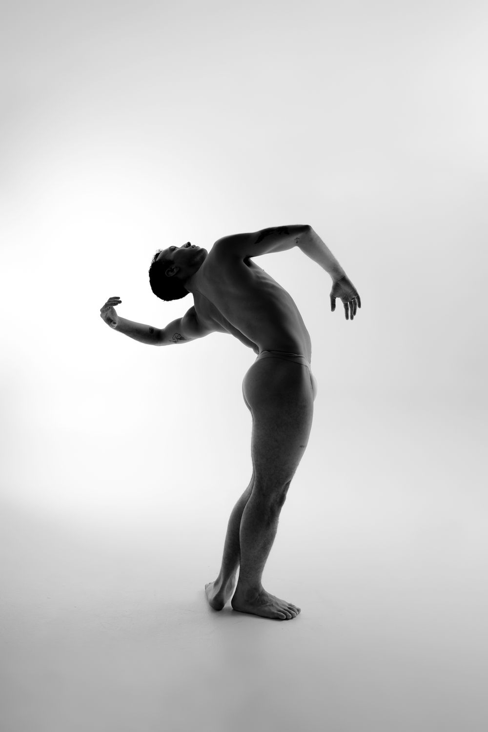 Black and white artistic nude silhouette of a dancer arching backwards in an elegant pose against light background.